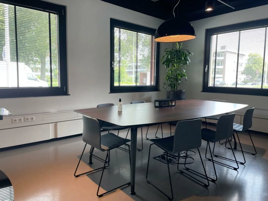 Vergaderruimte aan de De Cuserstraat met grote tafel, zes stoelen, kamerplant en hanglamp bij drie ramen met jaloezieën.